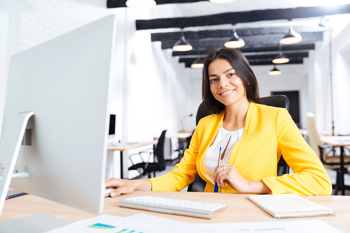 A smiling woman in a yellow blazer sits at a desk in a modern office, holding a pen, with a computer and notebook in front of her.