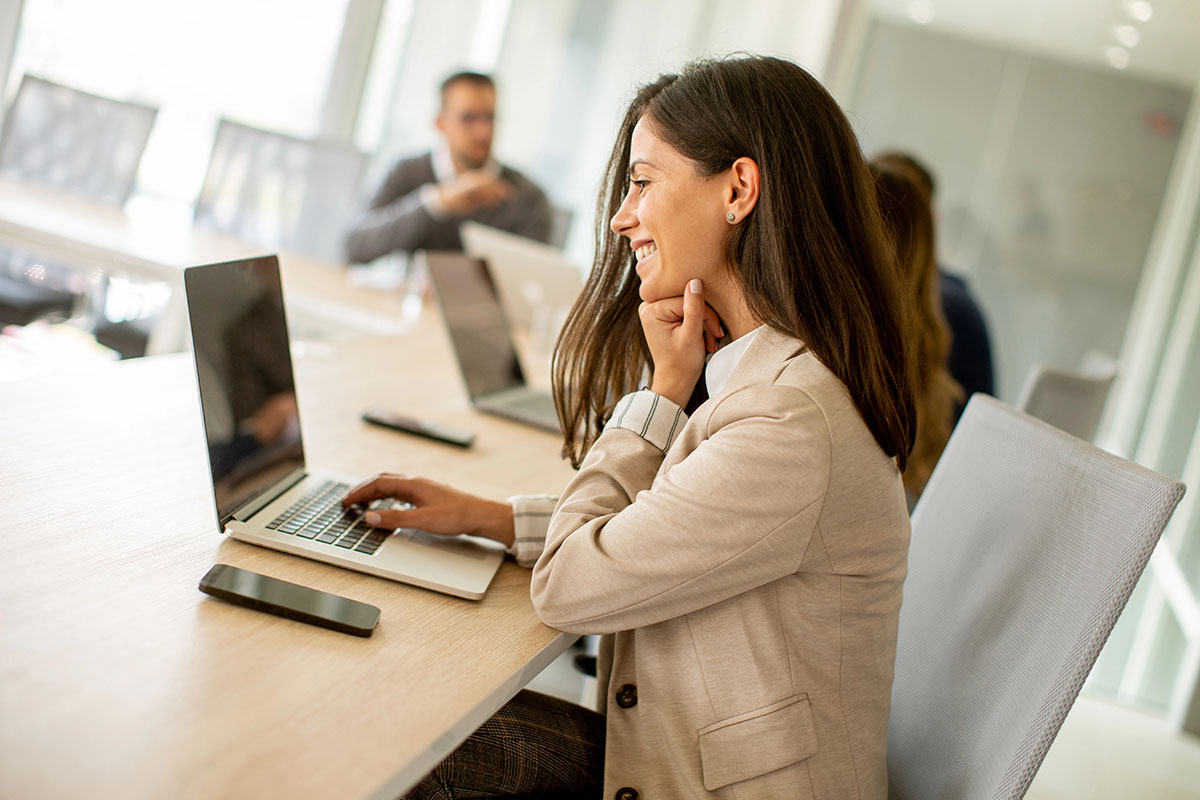 A woman in a beige blazer smiles as she works on a laptop at a conference table, with several colleagues engaged in discussion in the background.