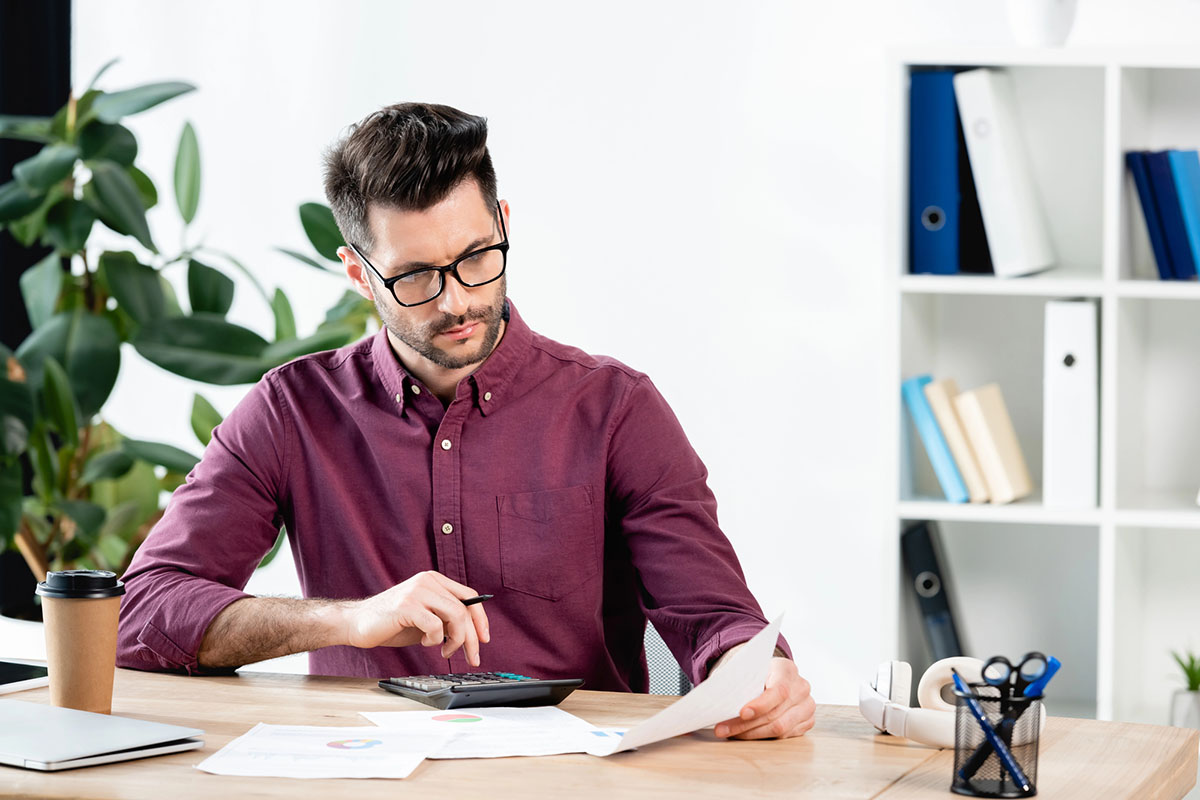 A young man in a maroon shirt and glasses sits at a desk, reviewing documents while using a calculator. A coffee cup and a laptop are on the desk, along with a container of pens and folders in the background.