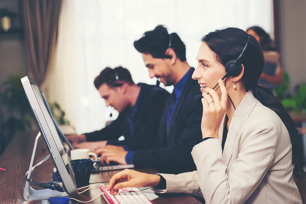 A diverse group of three people working at computers in a modern office setting. The woman in the foreground, wearing a blazer and a headset, smiles while speaking into the microphone. Two men in the background, also wearing headsets, are focused on their screens. Natural light filters through a window, and there's greenery visible in the background.