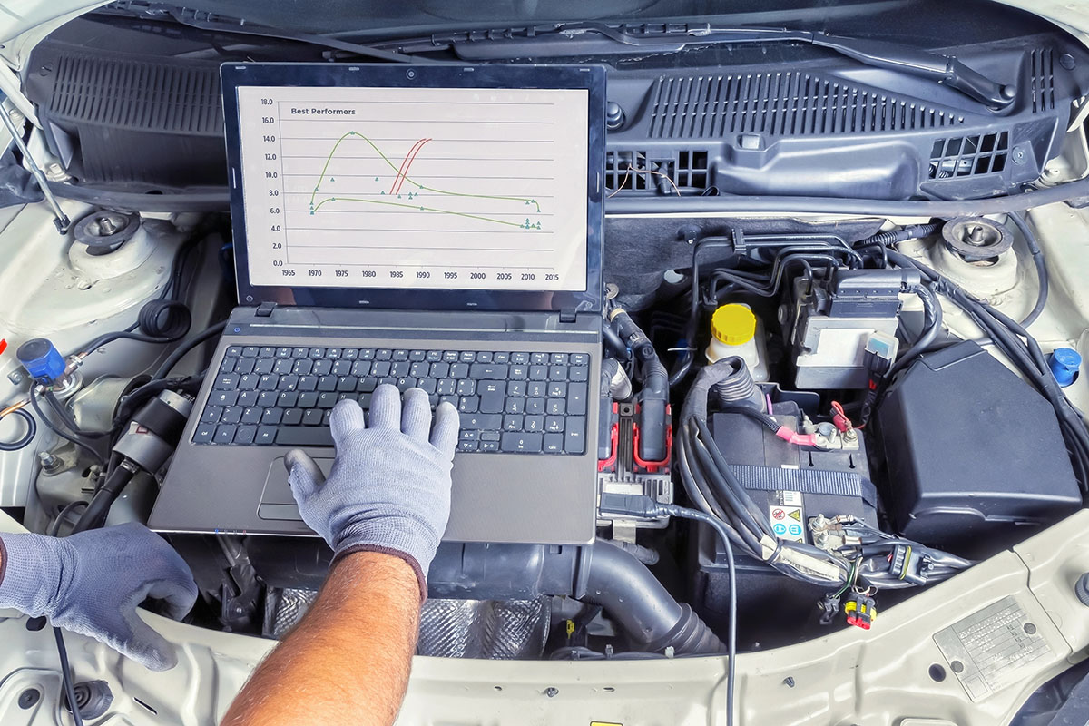 A mechanic's hands, wearing gray gloves, operate a laptop placed on the engine of a car, displaying a graph titled "Best Performers" with data points and trends over the years from 1970 to 2015. Various car components are visible around the laptop.