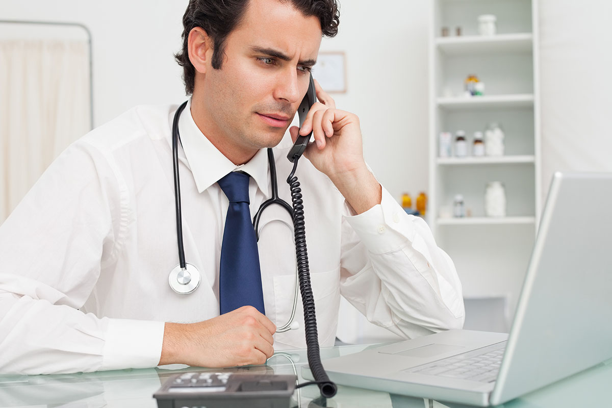 A man in a white shirt and tie with a stethoscope around his neck is talking on the phone while looking at a laptop in a professional office setting.