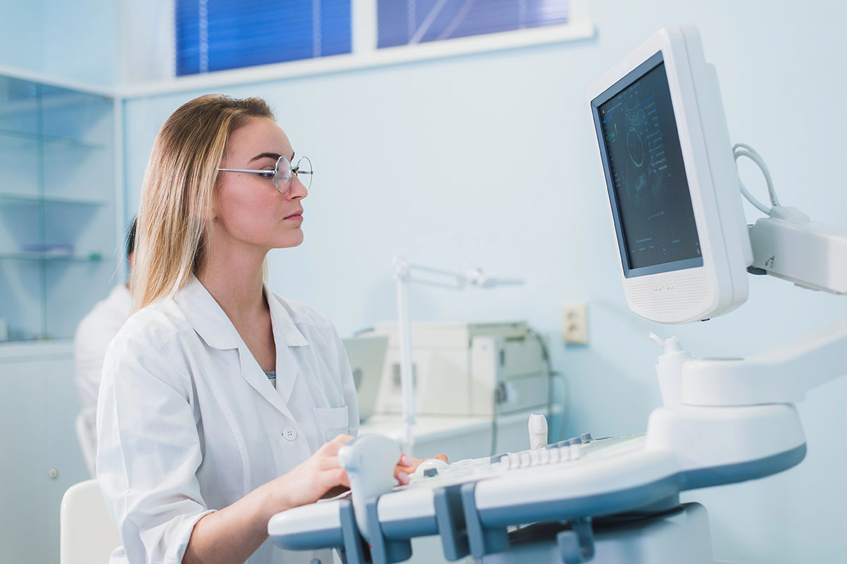 A female medical professional in a white lab coat and glasses focuses intently on a monitor displaying medical images, sitting at a diagnostic machine in a well-lit medical clinic.