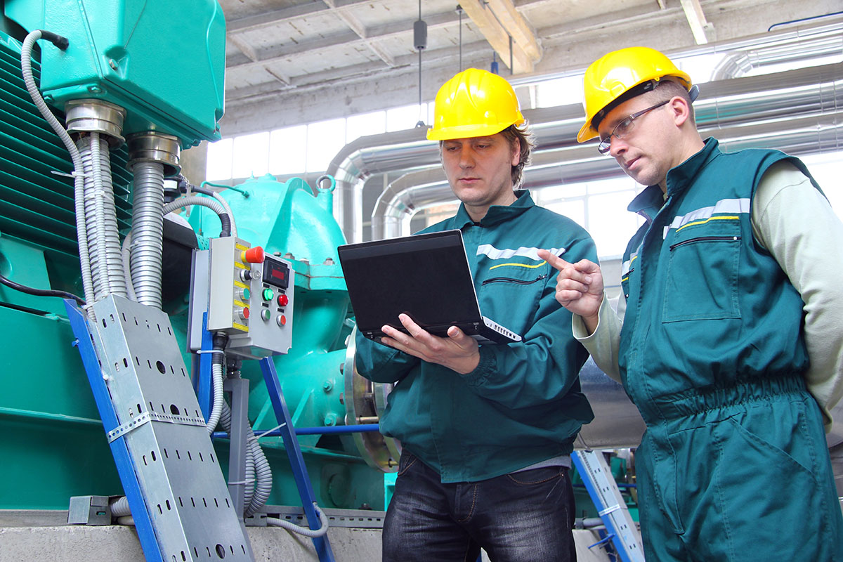 Two workers in hard hats are discussing information displayed on a laptop in an industrial setting, with machinery and control panels in the background.