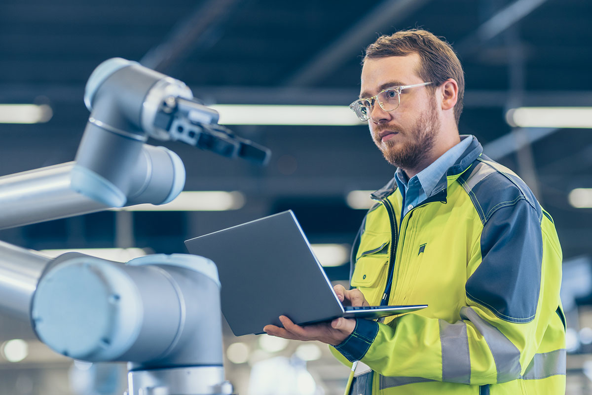 A man in a high-visibility jacket holds a laptop while interacting with a robotic arm in a modern industrial setting.