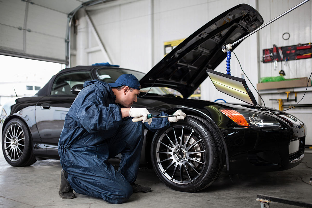 A mechanic wearing a blue jumpsuit and gloves kneels beside a black sports car, inspecting the tire while the hood is open and a work light is positioned above.