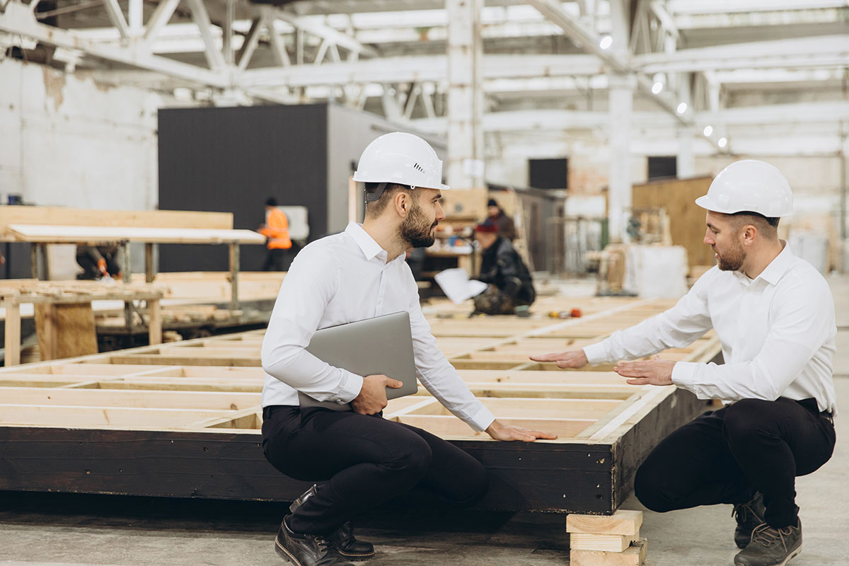 Two construction professionals in hard hats discuss plans while examining a wooden framework in a large, industrial workspace. One man is seated, holding a laptop, while the other gestures towards the structure. In the background, other workers are engaged in various tasks.