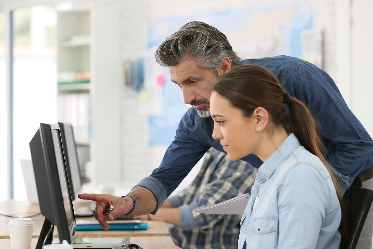 A man with gray hair leans over to assist a woman with a ponytail at a computer desk, both focused on the screen, while a colleague in the background observes.