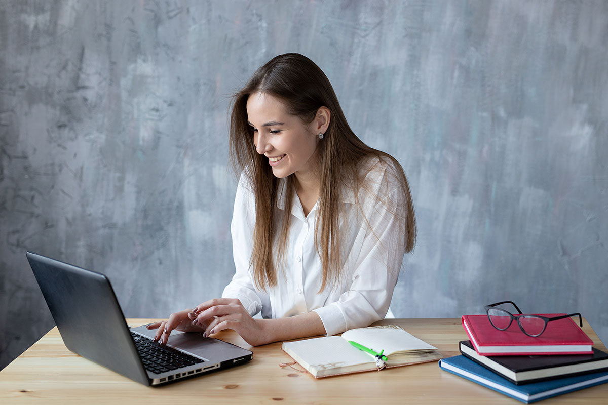 A smiling woman with long hair sits at a wooden table, focused on typing on a laptop. Nearby, there is a notebook with a green pen and a stack of books, while a pair of glasses rests on top of one of the books. The background features a textured gray wall.