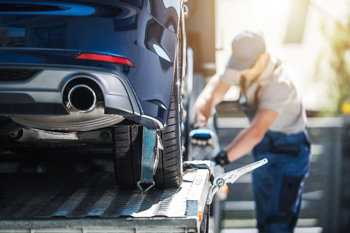 A blue car is being loaded onto a tow truck, with a worker securing it using a strap. The background is blurred, highlighting the car and the worker in a sunny outdoor setting.